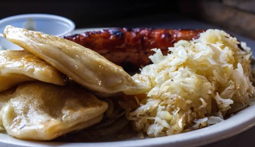 A plate of pierogi, sauerkraut, and a grilled sausage, served with a small cup of sauce on the side.