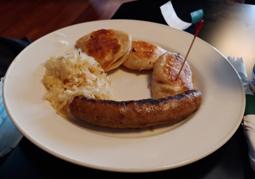 A plate with a grilled sausage, three potato dumplings, and a serving of sauerkraut.