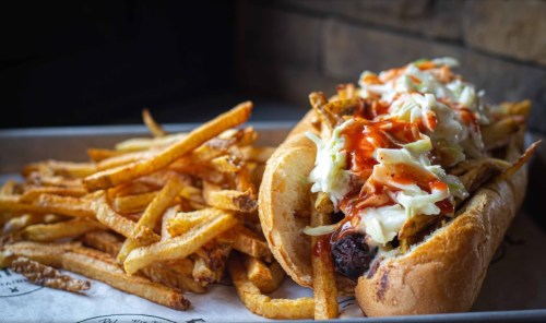 A loaded hot dog topped with coleslaw and sauce, served with a side of crispy French fries on a tray.