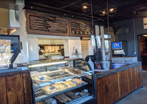 Interior of a restaurant featuring a display case with meats, a menu board, and a drink station.