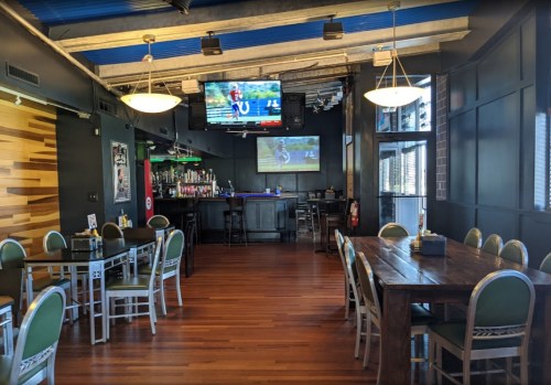 Interior of a restaurant with wooden floors, a bar area, and TVs showing sports. Tables and chairs are arranged for dining.