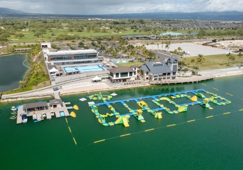 Aerial view of a water park with inflatable attractions, a pool, and a modern building by a lake surrounded by greenery.