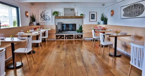 Bright restaurant interior with wooden floors, tables, and a decorative fireplace. Plants and artwork adorn the walls.
