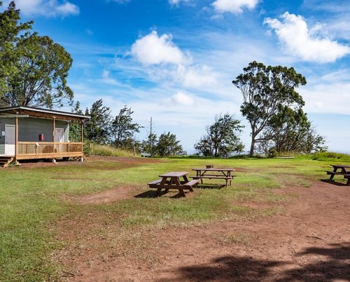 A grassy area with picnic tables and a small building, surrounded by trees under a blue sky with clouds.