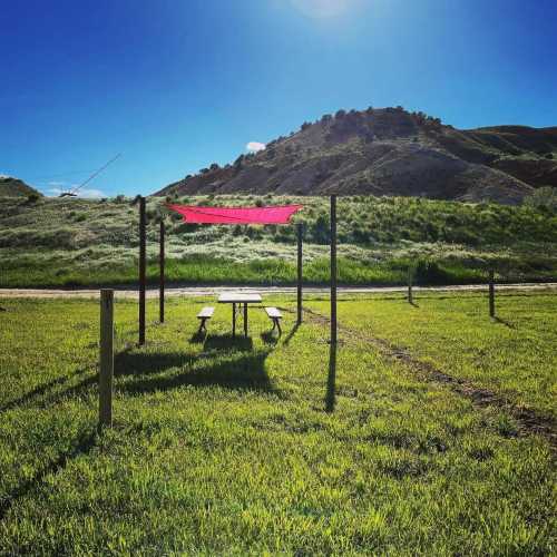 A picnic table under a red canopy in a grassy area, with hills and a clear blue sky in the background.