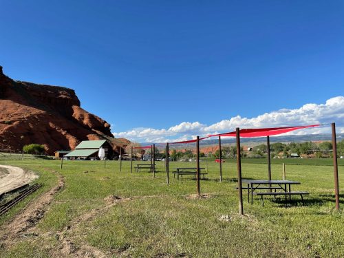 A grassy area with picnic tables and red canopies, surrounded by red rock formations and a clear blue sky.