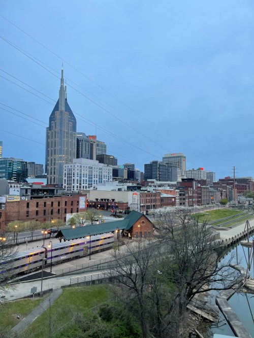 City skyline featuring a tall building with a spire, alongside a river and green space under a cloudy sky.