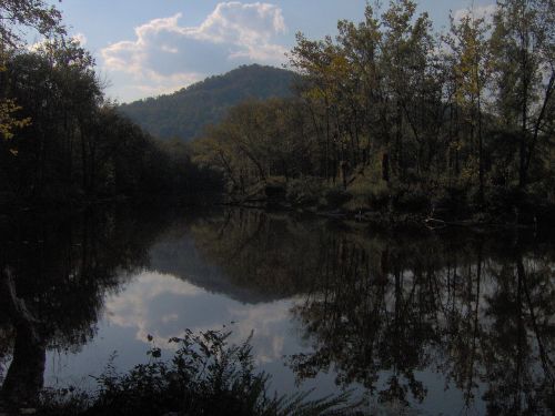 A serene river scene with trees and a mountain reflected in the calm water under a cloudy sky.