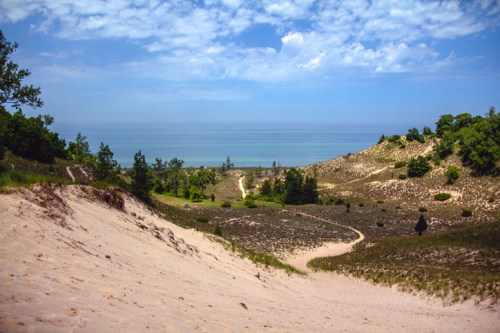 A scenic view of rolling sand dunes leading to a calm lake under a blue sky with scattered clouds.