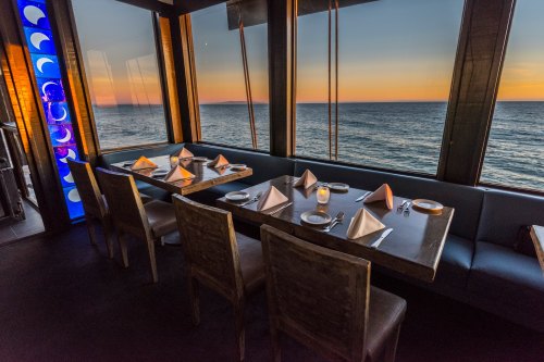 A seaside restaurant with a table set for dinner, featuring large windows showcasing a sunset over the ocean.