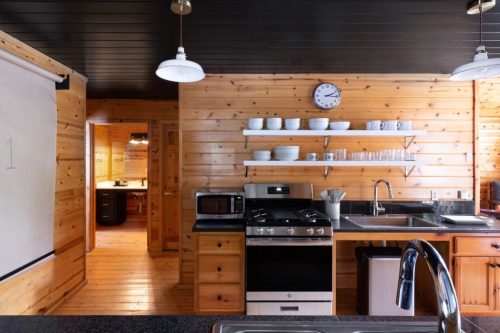 Modern kitchen with wooden walls, black countertops, a stove, and open shelving displaying dishes and glassware.