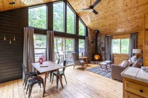 Cozy cabin interior with large windows, wooden walls, a fireplace, and a dining area. Natural light fills the space.