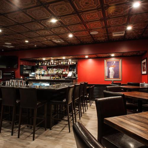 A stylish bar with a dark wood counter, high chairs, and a red wall adorned with framed art and shelves of liquor.