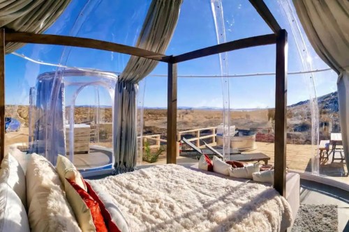 A cozy, transparent dome bedroom with a plush bed, offering stunning desert views and outdoor seating.