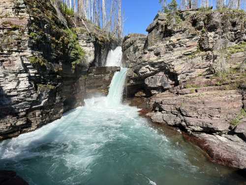 A waterfall cascades into a turquoise pool surrounded by rocky cliffs and sparse trees under a clear blue sky.