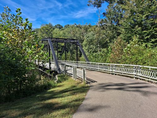 A metal bridge spans a path surrounded by lush greenery under a clear blue sky.