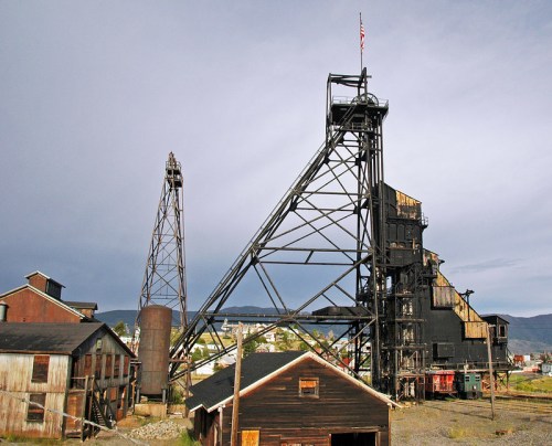 Historic mining structures with a tall headframe and buildings under a cloudy sky. An American flag flies atop.