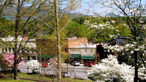 A scenic view of a small town with blooming trees, shops, and mountains in the background on a sunny day.