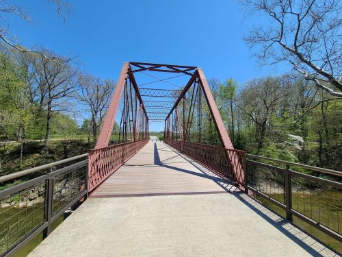 A red metal bridge spans a river, surrounded by trees under a clear blue sky.