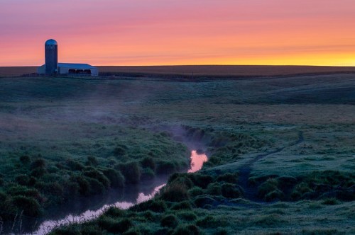 A serene farm scene at sunrise, featuring a barn, silo, and a misty stream surrounded by lush green fields.