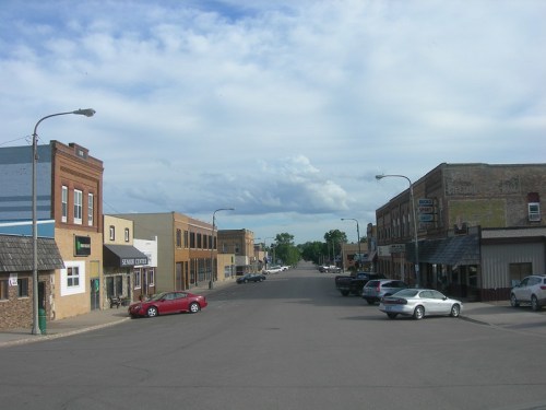 A quiet street lined with small businesses and parked cars under a cloudy sky.