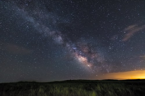 A stunning night sky filled with stars and the Milky Way, set against a dark landscape with a hint of orange glow.