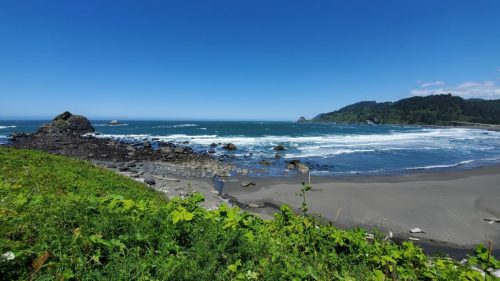 A scenic coastal view featuring waves crashing on rocky shores under a clear blue sky and lush green hills.