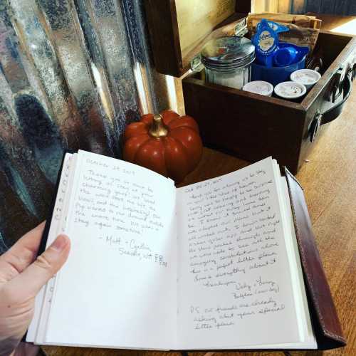 A hand holds an open notebook with handwritten notes, next to a small pumpkin and a wooden box filled with jars.