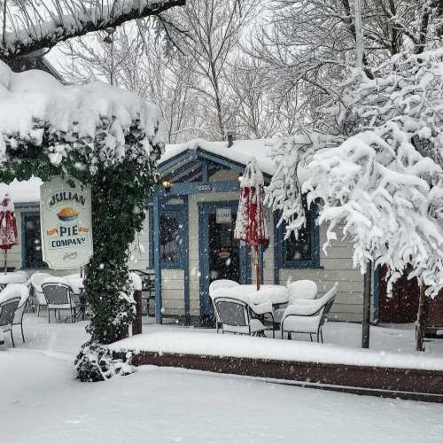 A cozy pie shop covered in snow, with outdoor seating and festive umbrellas, surrounded by winter trees.