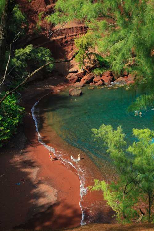 A scenic view of a red sand beach with turquoise water, surrounded by lush greenery and rocky cliffs.