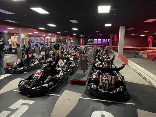 A group of go-kart racers in black karts, ready to race on an indoor track with colorful lights in the background.