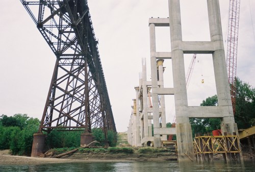 A view of a river with a steel bridge on the left and concrete pillars under construction on the right.