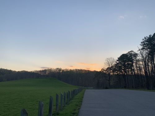 A serene landscape at dusk, featuring a grassy hill, a tree line, and a quiet road under a colorful sky.
