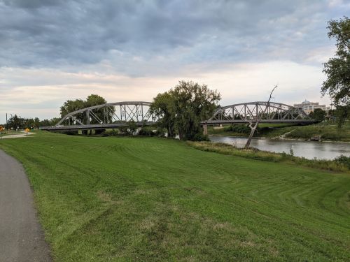 A metal bridge spans a river, surrounded by green grass and trees under a cloudy sky.