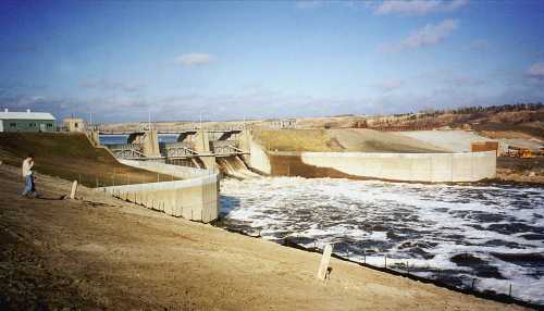 A large dam with water flowing over it, surrounded by grassy land and a clear blue sky. A person walks nearby.