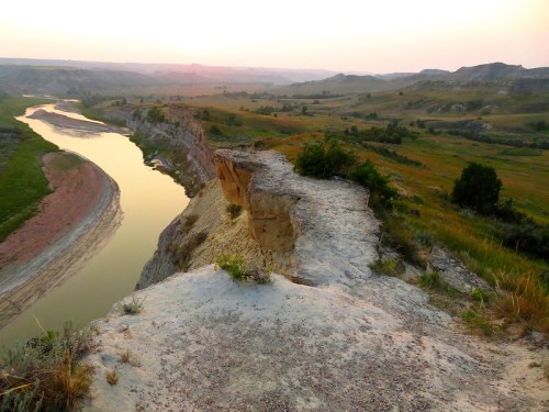 A scenic view of a river winding through a valley at sunset, with cliffs and lush greenery in the foreground.