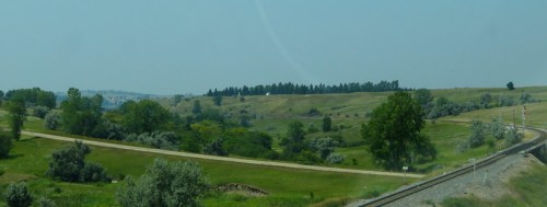 A scenic view of rolling green hills, trees, and a dirt road alongside a railway track under a clear sky.