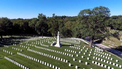Aerial view of a cemetery with rows of white headstones and a central monument surrounded by trees.