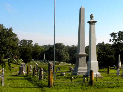 Two tall stone monuments in a green cemetery, surrounded by trees and a clear blue sky.