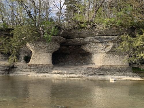 A rocky cliff with two large, rounded formations above a calm river, surrounded by trees and greenery.