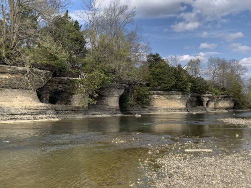 A serene riverbank with layered rock formations and trees, under a partly cloudy sky.