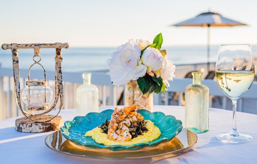 A beautifully plated shrimp dish on a turquoise plate, with a glass of white wine and flowers, set by the ocean.