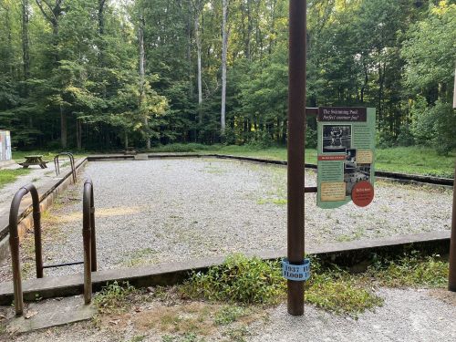 A gravel area surrounded by trees, featuring a sign about a pet agility course and a metal railing.