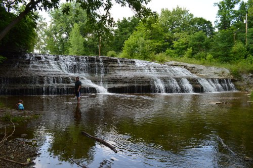 A man wades in shallow water near a waterfall, with a child sitting on the bank surrounded by lush greenery.
