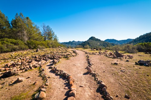 A winding dirt path surrounded by rocky formations and greenery, leading through a mountainous landscape under a clear blue sky.