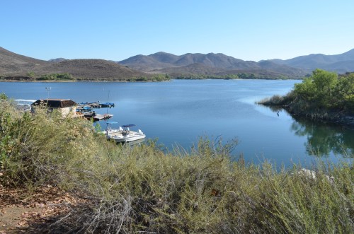 A serene lake surrounded by mountains, with boats docked and lush greenery along the shore under a clear blue sky.