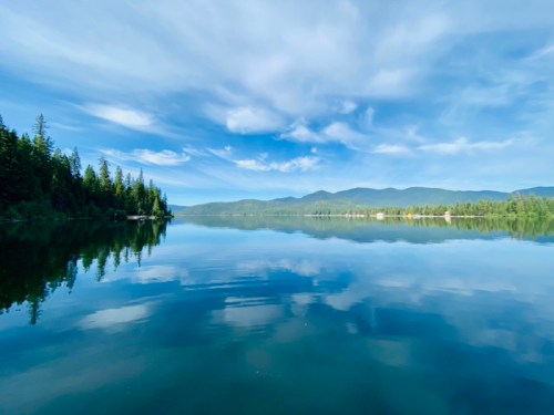 A serene lake reflecting blue skies and mountains, surrounded by lush green trees.