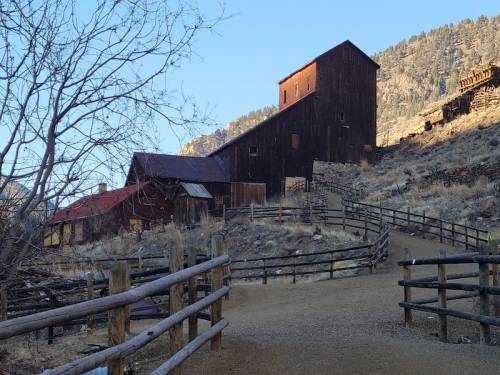 A rustic wooden building on a hillside, surrounded by trees and a dirt path, with mountains in the background.