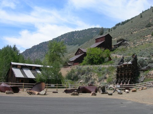 Historic mining buildings on a hillside, surrounded by trees and rocky terrain under a blue sky with scattered clouds.