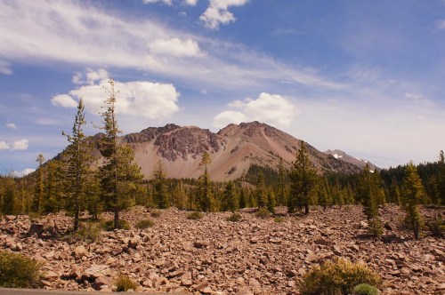 A rocky landscape with scattered trees in the foreground and a mountain under a blue sky with clouds in the background.